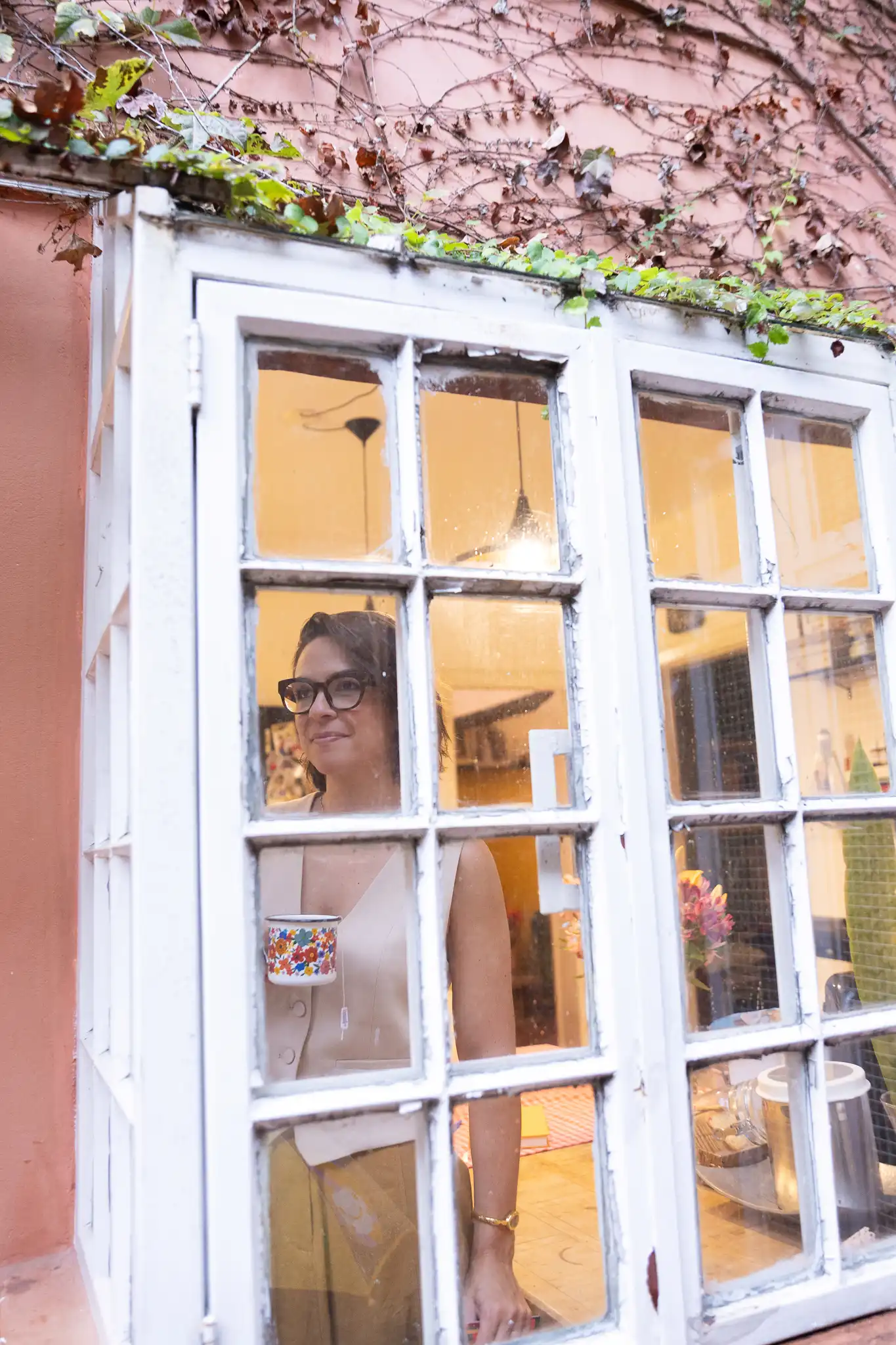 Nicole vista pela janela de casa, com uma caneca de flores na mão